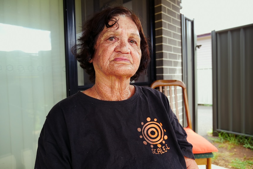 Woman sits in black t-shirt with Aboriginal art logo, in front of grey brick house with glass door.