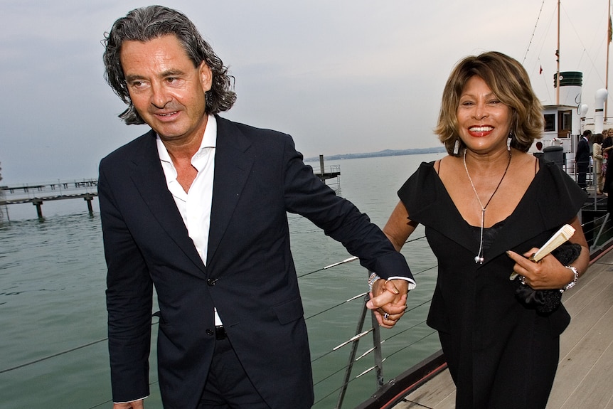 A man in a suit with no tie leads Tina Turner, wearing a black dress and smiling.