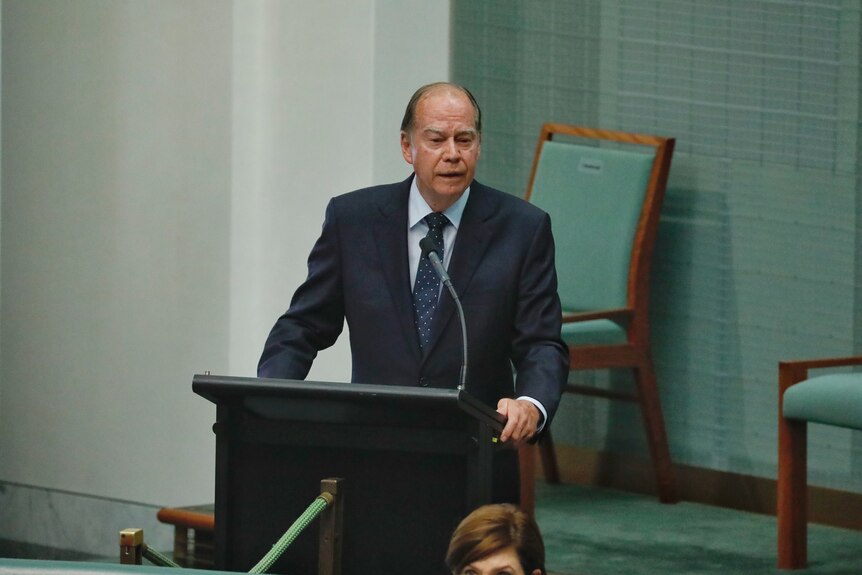 An older man in a suit stands before a lectern in the House of Representatives chamber.