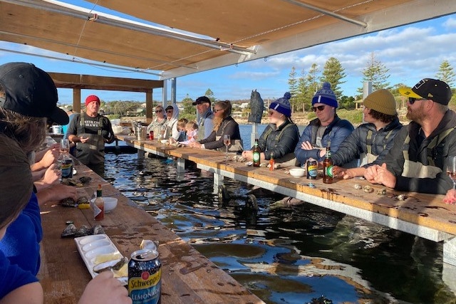 People sit around a floating pontoon wearing beanies with drinks and oysters in their hands