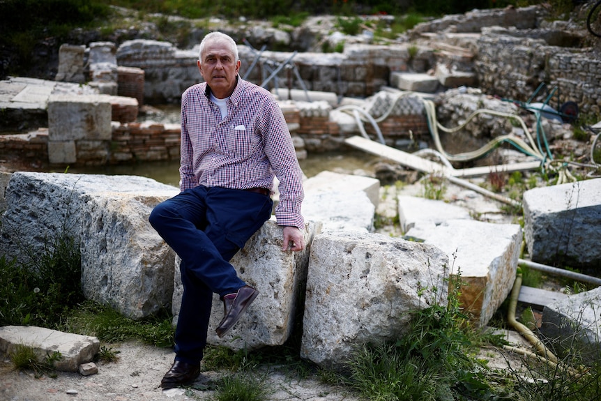 An older white man in a pink and purple checked shirt sits on a low stone wall amid archaeological ruins.