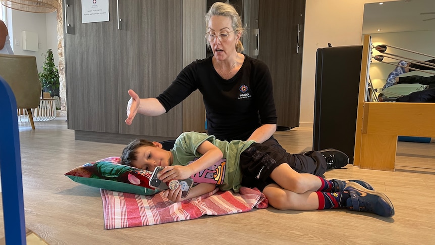 A child holds a phone lying on his side whilst the physio moves him into a position better for posture 