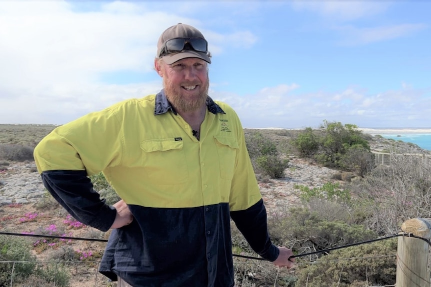 Man in high vis, hand leaning on fence, hat and sunnies on head, in front of low scrub and ocean in background 