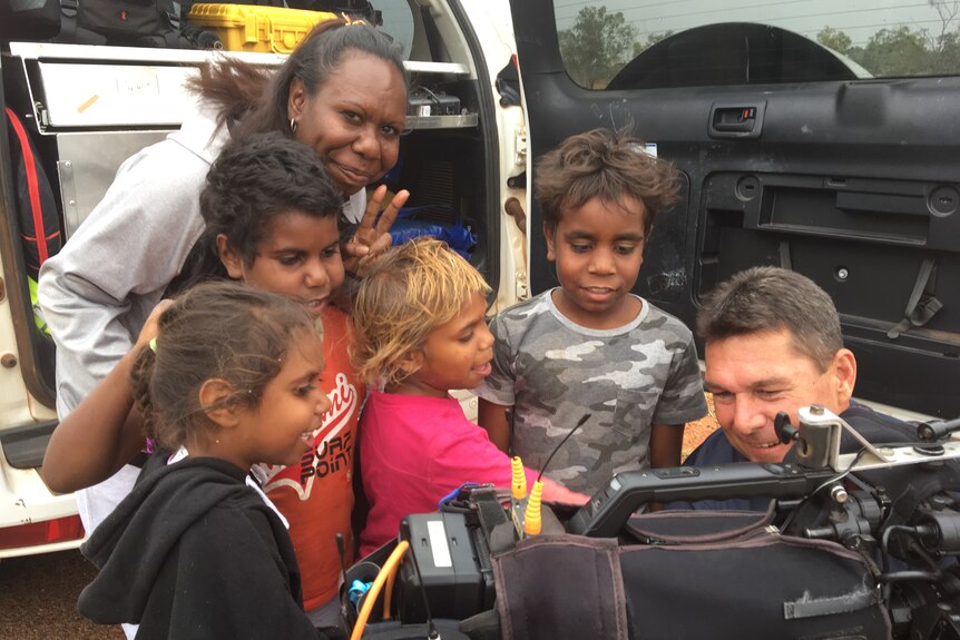 A group of children flock around a cameraman and his camera, the man is smiling.