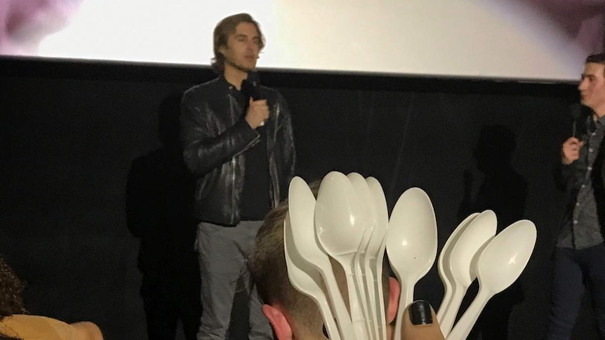 Greg Sestero on stage talking with plastic spoons held in foreground