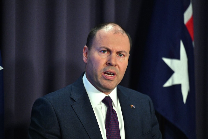 Josh Frydenberg making a speech, wearing a suit with an Australian flag while standing in front of an Australian flag.