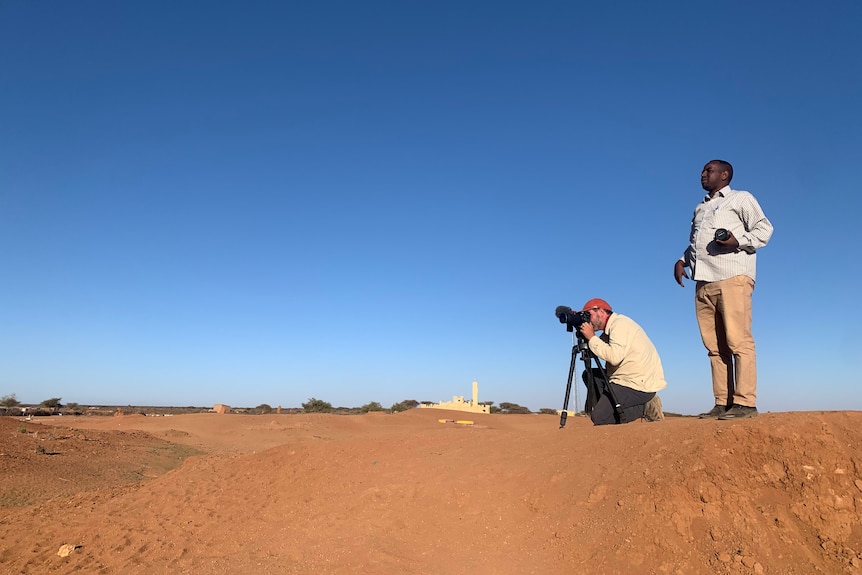 Cameraman films a dry, desert landscape as another man stands behind him watching.