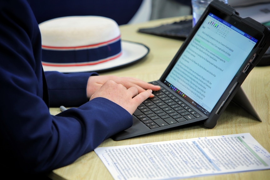 A school student wearing a uniform with long royal blue sleeves types on a tablet computer.