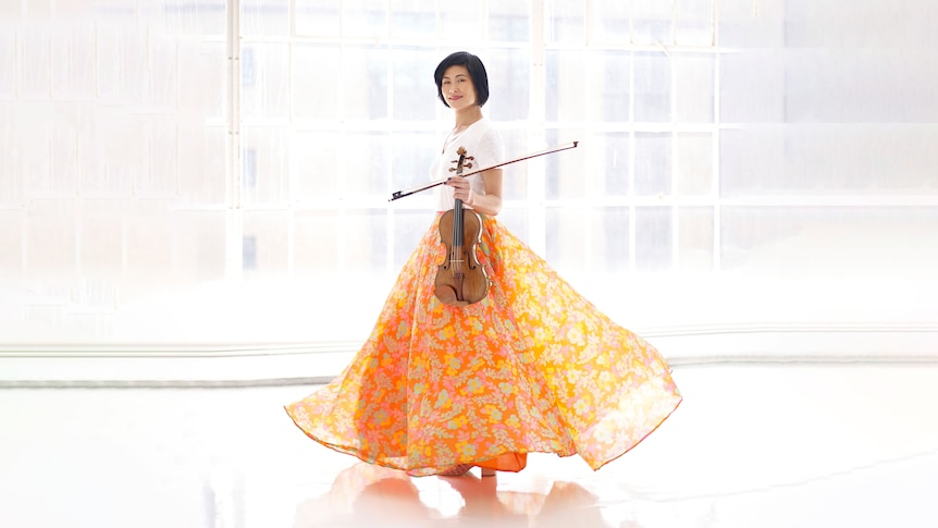 Violinist Jennifer Koh stands in a white room holding a violin, she is wearing an orange skirt and a white top.