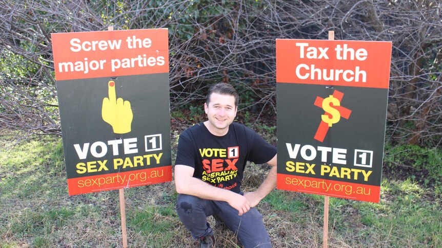 Steven Bailey poses with signs saying 'screw the major parties', and 'tax the church'.