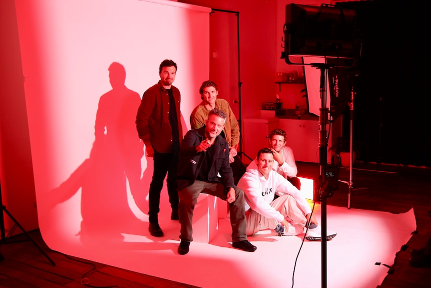Five men pose against a white backdrop screen for a photoshoot, illuminated by red shadow lighting