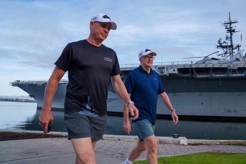 Mark Hammond and Anthony Albanese wearing exercise clothes walk past a large warship
