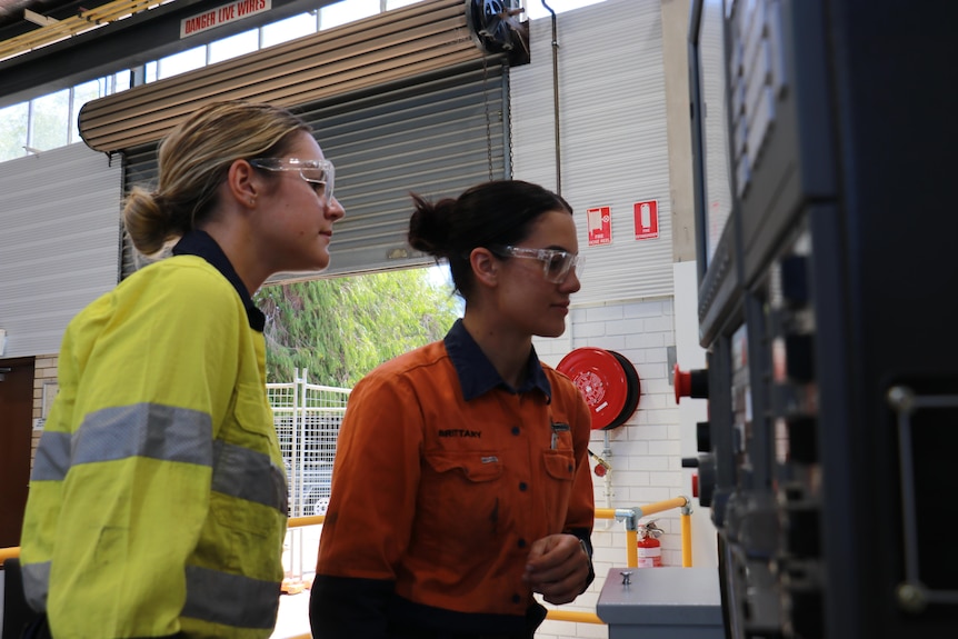 Two young women, one wearing orange high-vis wear and the other yellow, in safety glasses looking at a mechanical panel