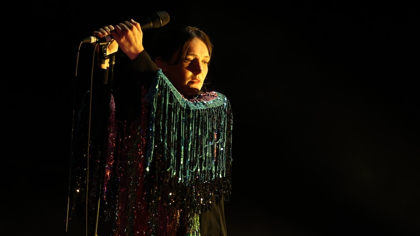 Sarah Blasko holds a microphone aloft on stage. She wears a green tasseled jacket.