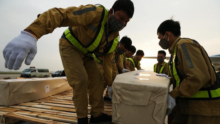 From a low angle, you view a group of airport staff lifting a fabric-wrapped coffin from a wooden freight crate on a runway.