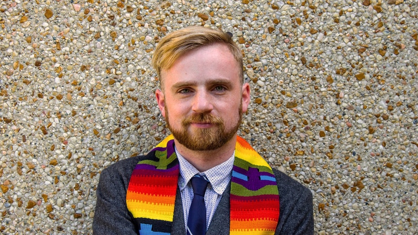 Man wearing suit and a rainbow-coloured stole over his shoulders.