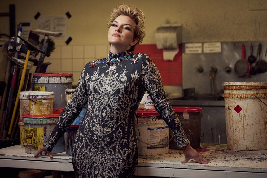 A blonde, short-haired woman, wearing an extravagant dress, leans back on a workshop bench, with a defiant expression