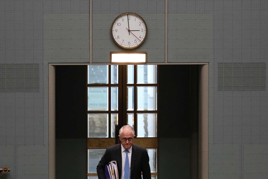 Malcolm Turnbull, frowning, walks through the House of Representatives door. A clock on the wall above his head reads 2:59.