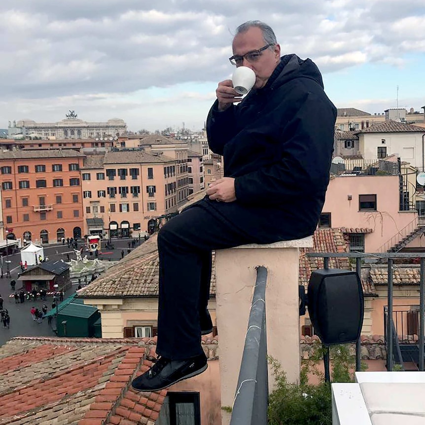 Antonis Mavropoulos, a grey-haired Greek man with glasses, sits on a balcony ledge and drinks a coffee.