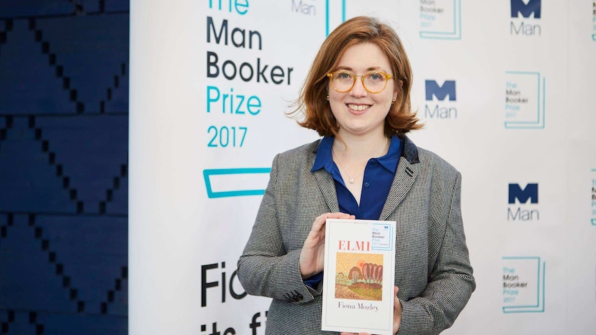 Colour photograph of novelist Fiona Mozley holding her shortlisted book in front of a media wall.