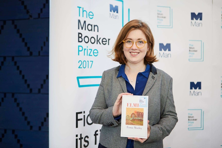 Colour photograph of novelist Fiona Mozley holding her shortlisted book in front of a media wall.