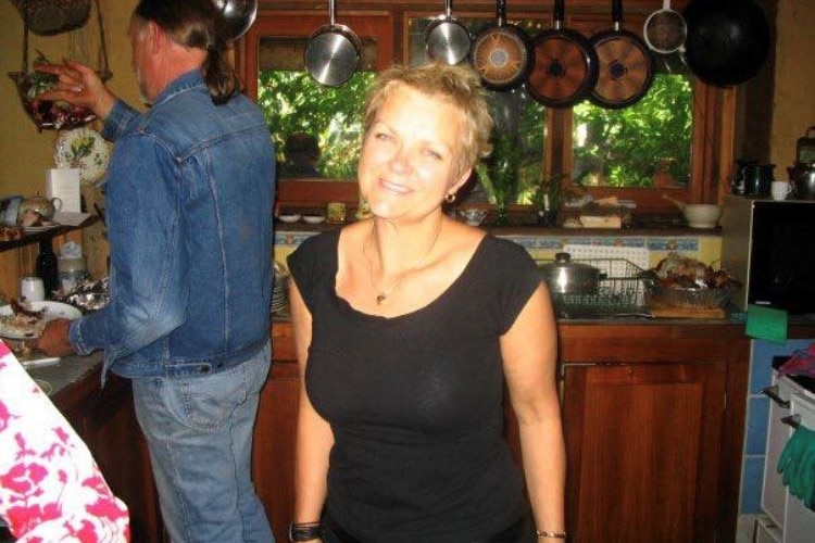 A woman with short blonde hair, wearing a black shirt, stands smiling in a kitchen.