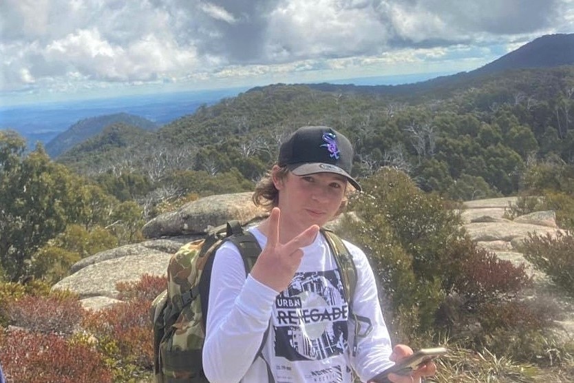 A boy in his early teen years gives the camera a peace sign, standing on a rocky outcrop.