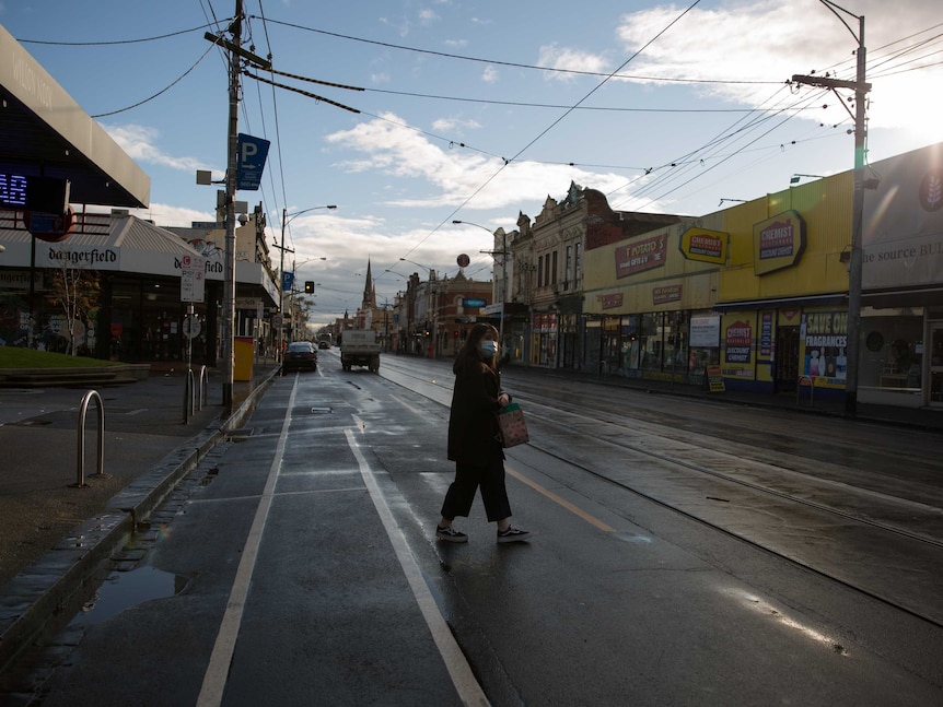 A woman wearing a mask crosses the wet road in front of a kebab shop and chemist as the sun beams