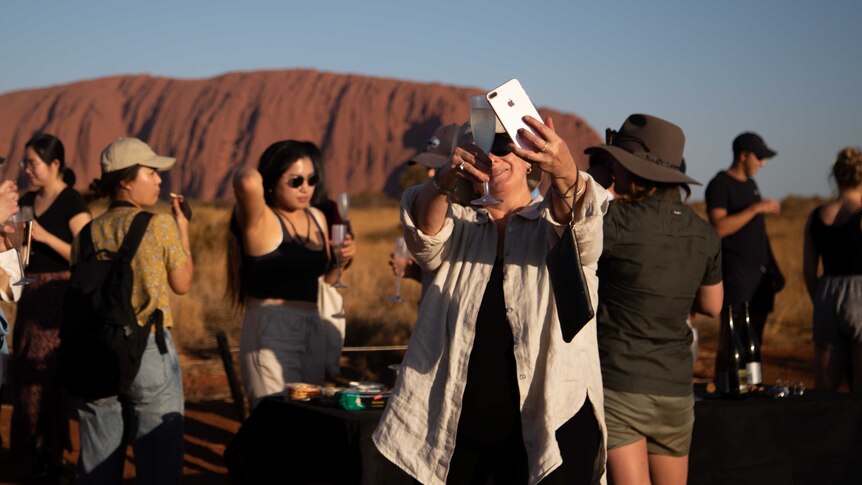 A traveller takes a photo of her champagne with Uluru behind her. There is a large crowd of people in the area.