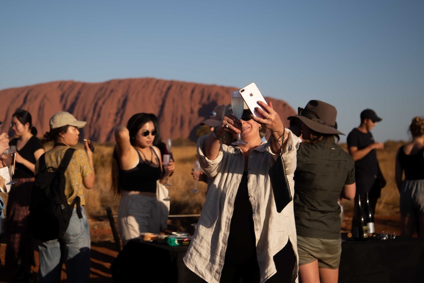 A traveller takes a photo of her champagne with Uluru behind her. There is a large crowd of people in the area.