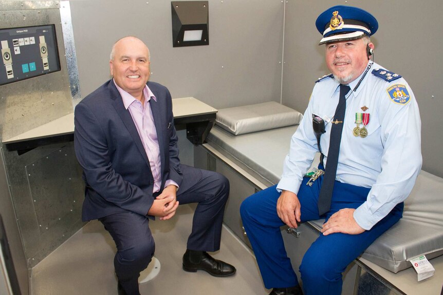 Minister left sits on chair and gaol governor sits on bed inside new 'rapid-build' cell.