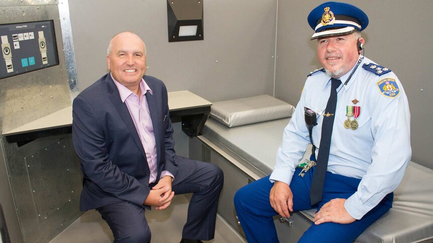 Minister left sits on chair and gaol governor sits on bed inside new 'rapid-build' cell.