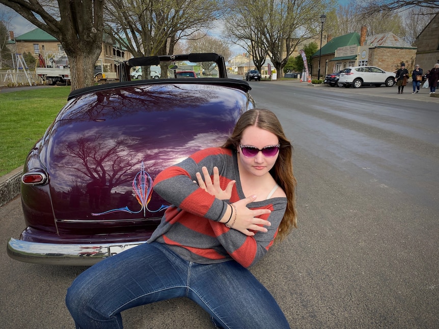 A young woman with long hair and sunglasses, crouched down with crossed arms, posing in front of a purple vintage car.