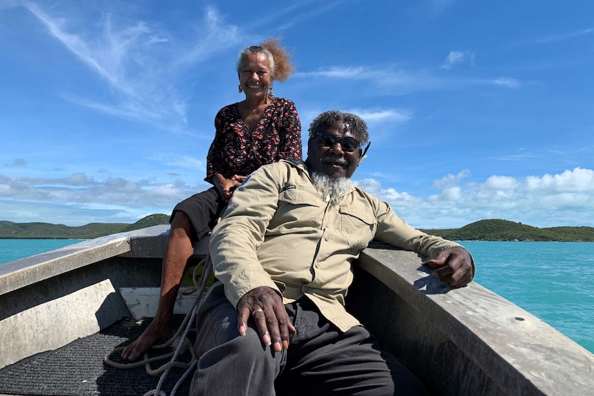 Enid Tom and Milton Savage sit on a dinghy surrounded by clear blue water. Behind them in the distance is an island.