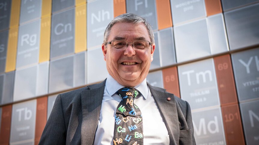A university professor with grey hair and glasses, smiling at the camera standing in front of a large periodic table.