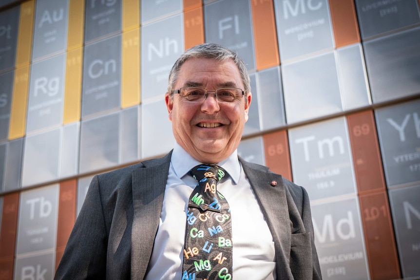 A university professor with grey hair and glasses, smiling at the camera standing in front of a large periodic table.