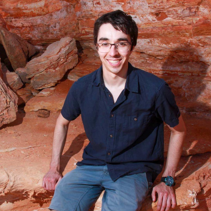 A dark haired young man sitting on a shelf of red rock, with more rock behind him.