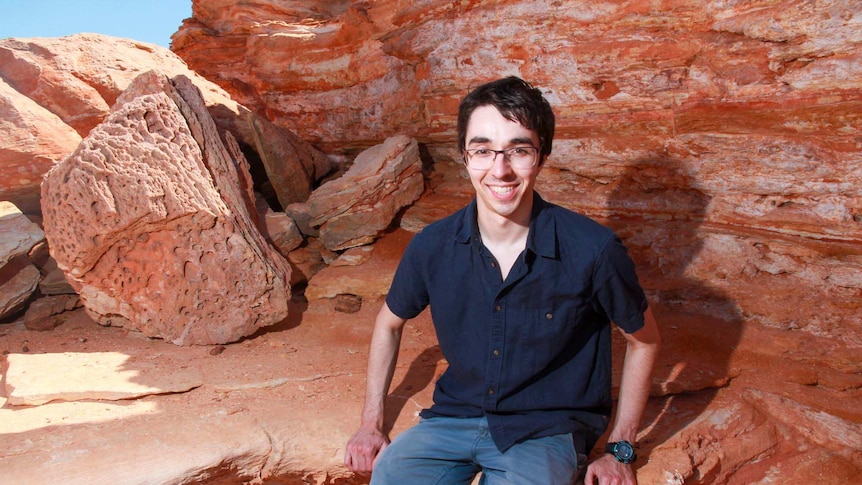 A dark haired young man sitting on a shelf of red rock, with more rock behind him.