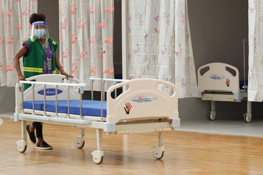 A woman wearing a mask and green uniform wheels a hospital bed in a room.