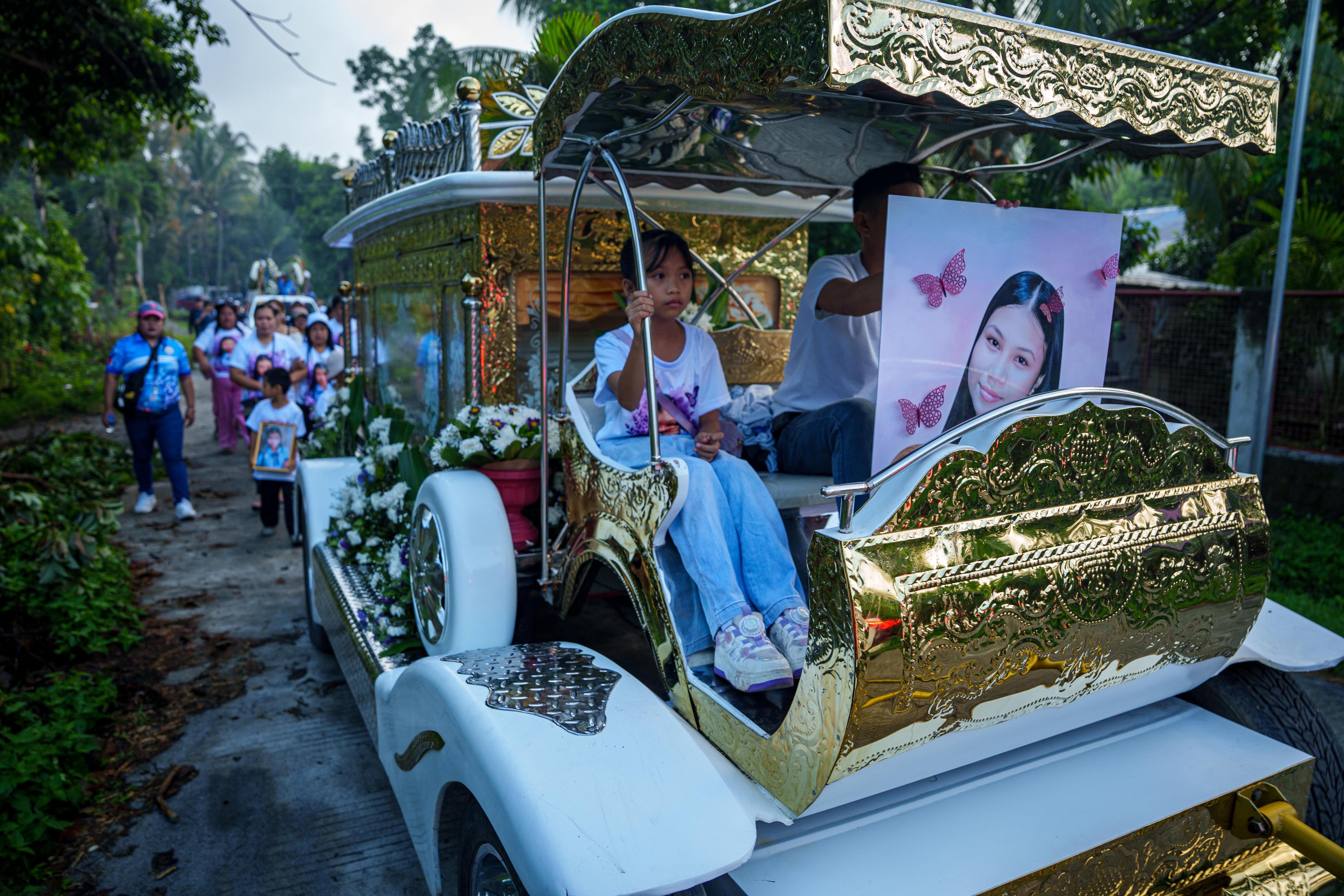 A golden carriage carrying a white coffin is driven along a dirt road with a group of people walking behind it.