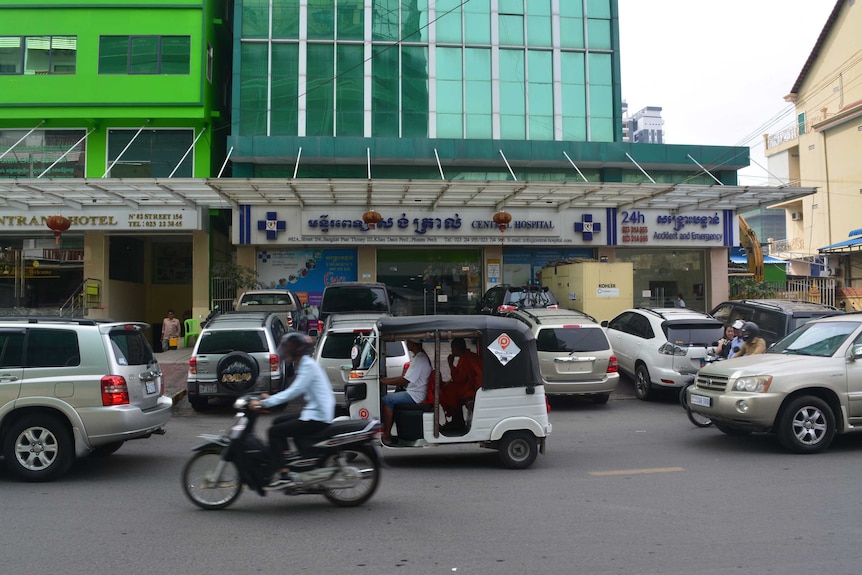 A tuk tuk and a motorbike drive past on a street with a large glass hospital building behind it.