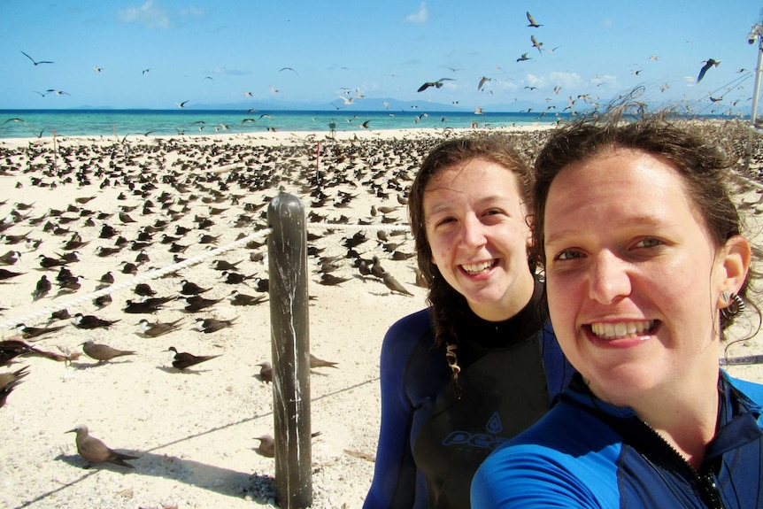 Two women smiling standing in front of hundreds of birds on a beach.