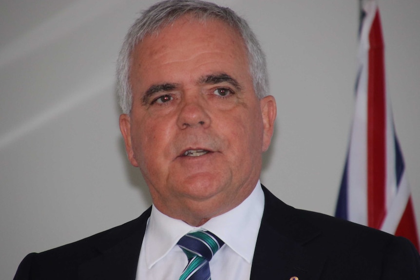 A head and shoulders shot of a man in a suit with an Australian flag behind him.