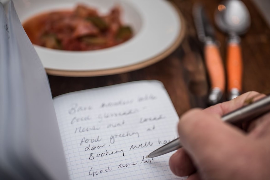 A hand holds a pen and makes notes in a note book next to a plate of food. The words 'good wine list' can be distinguished.