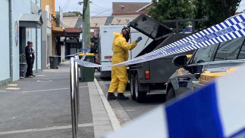 A crime scene officer in a yellow jumpsuit and face mask puts something into a vehicle outside Wellbeing Planet on a grey day.
