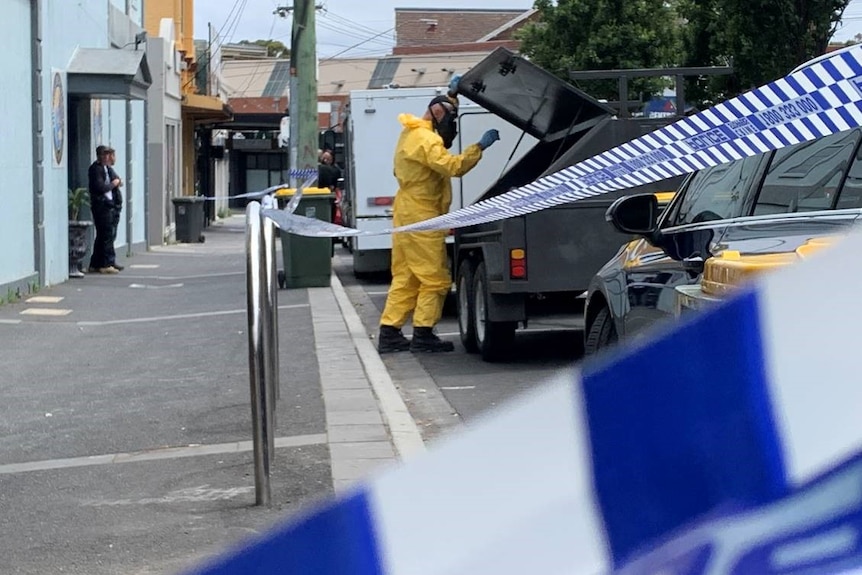 A crime scene officer in a yellow jumpsuit and face mask puts something into a vehicle outside Wellbeing Planet on a grey day.
