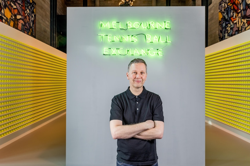 A man stands in front of rows of tennis balls and a neon sign reading 'Melbourne Tennis Ball Exchange'