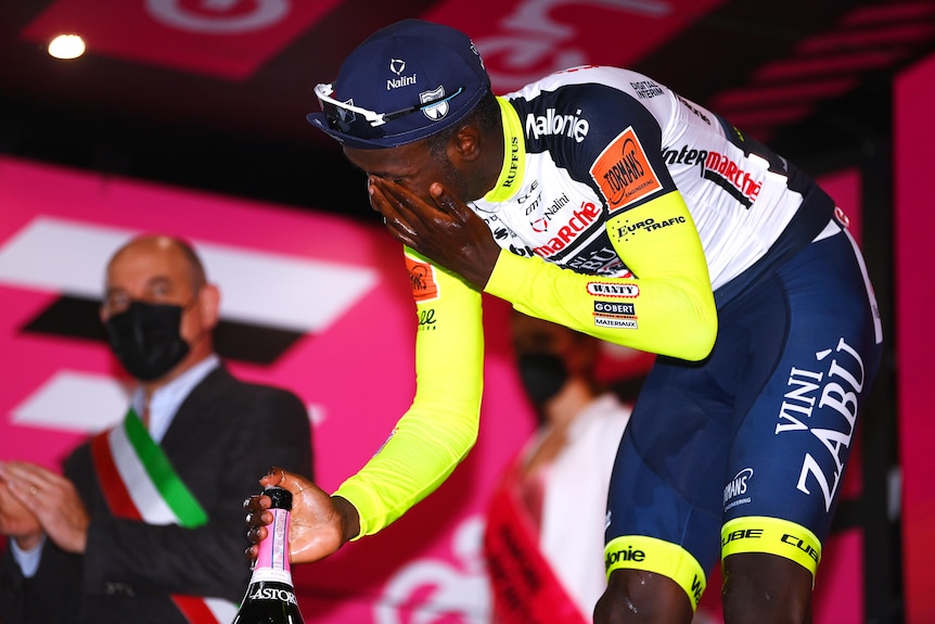 An African cyclist holds a champagne bottle in one hand and has his other hand over his eye. 
