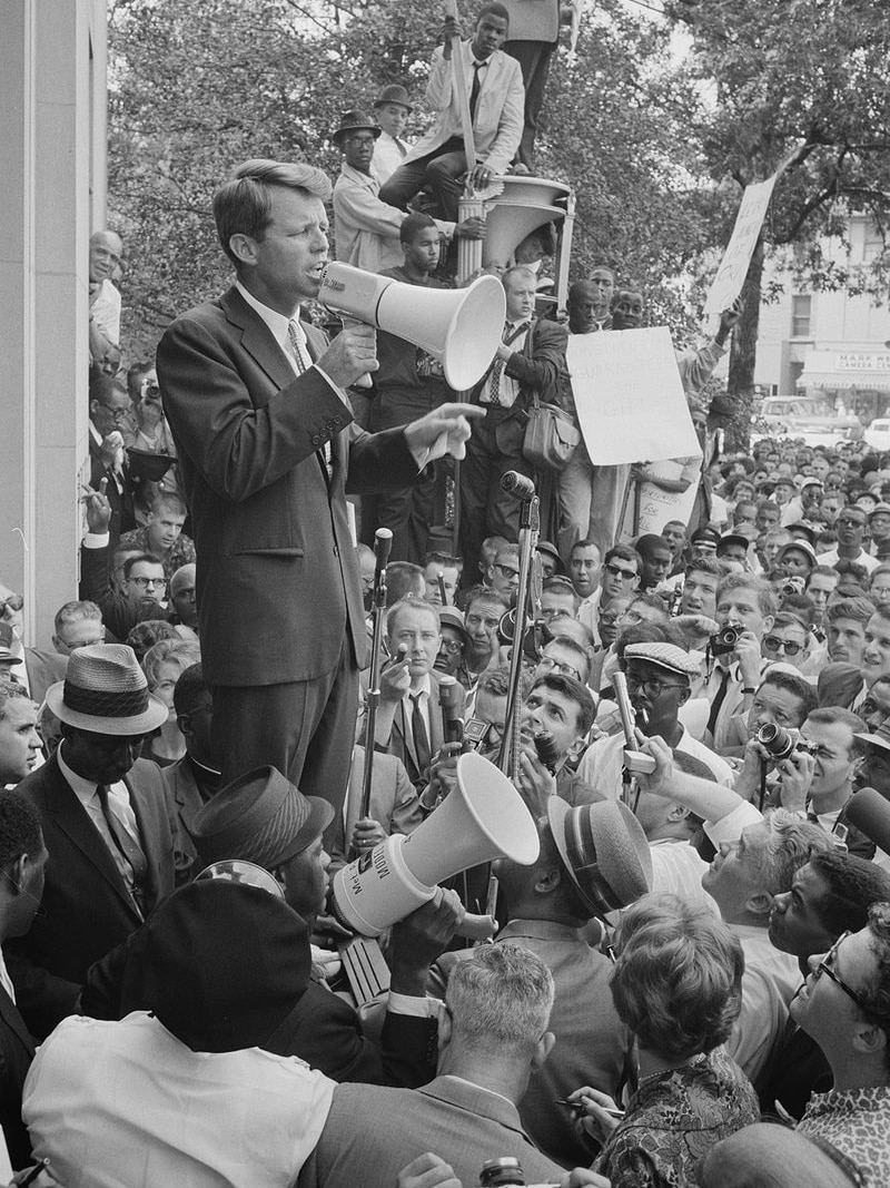 Bobby Kennedy stands on a podium with a megaphone, he is wrapped by a large crowd