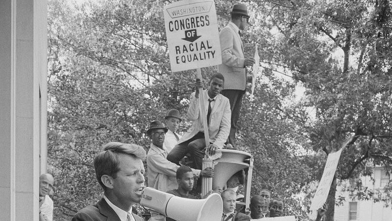 Bobby Kennedy stands on a podium with a megaphone, he is wrapped by a large crowd